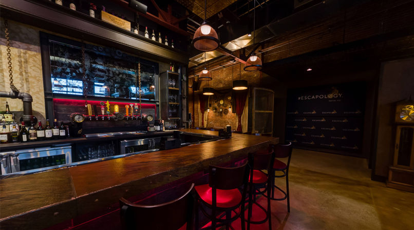 Moody industrial-style bar interior with a long wooden counter, row of red-cushioned bar stools, beer taps and bottles behind the bar, exposed brick walls, warm pendant lighting and vintage decor.