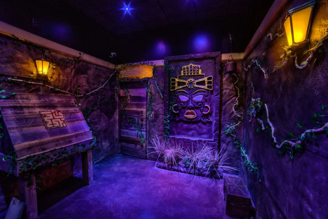 Purple-lit indoor Aztec-style escape room set with carved stone mask relief, vine-covered walls, glowing lanterns, wooden puzzle lectern and small treasure crate.