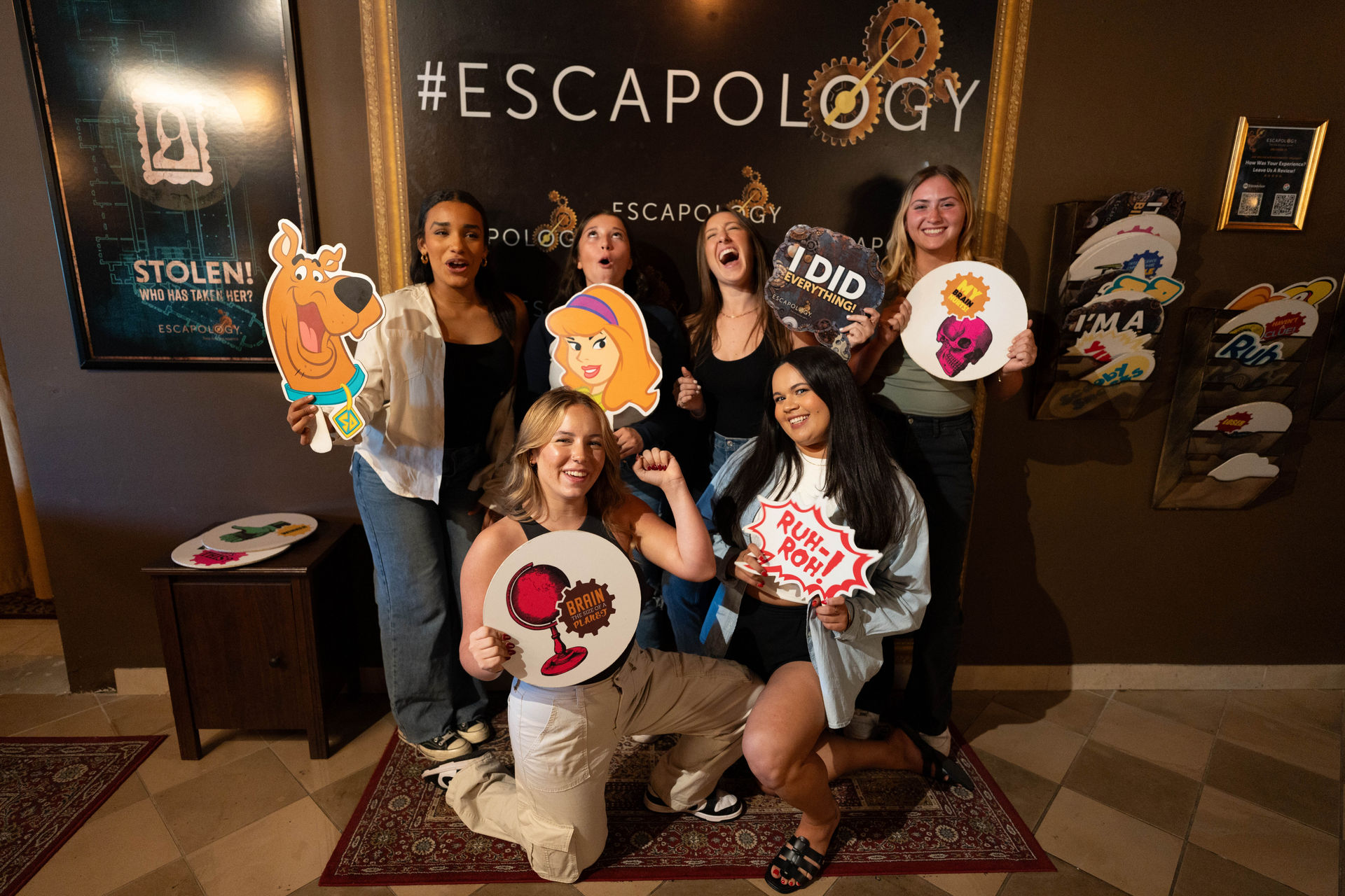 Six friends posing in an indoor escape-room photo area holding cartoon props like Scooby-Doo and Daphne cutouts and playful speech-bubble signs, celebrating a completed game.