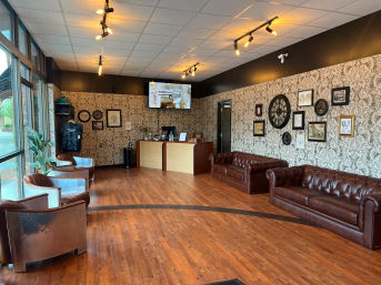 Sunlit reception/waiting room with floor-to-ceiling windows, hardwood floors, two brown leather Chesterfield sofas, riveted metal club chairs, a wooden reception desk, patterned wallpaper gallery wall and warm track lighting.