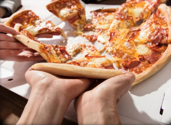 Hands grabbing cheesy pepperoni pizza slices from an open takeout pizza box on a table, melted cheese stretching as slices are pulled.