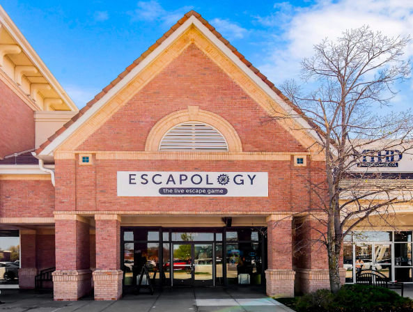 Brick gabled storefront in a suburban shopping plaza housing an escape-room venue, with a white sign above glass double doors, a leafless tree at the entrance, and a bright blue sky.
