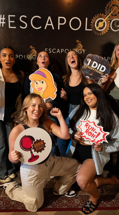 Six friends striking playful victory poses in an escape-room photo booth, holding cartoon character cutouts and speech-bubble props and smiling for a celebratory group shot.