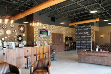 Industrial-chic escape room lobby with wooden reception counter, leather sofas, vintage wall clocks, patterned wallpaper, and exposed ductwork.