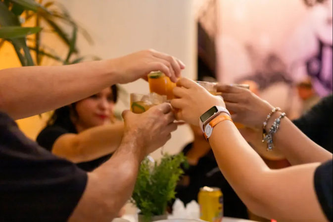 People clinking citrus cocktails in a lively indoor toast at a casual gathering, glasses with lime garnish and a small table plant.