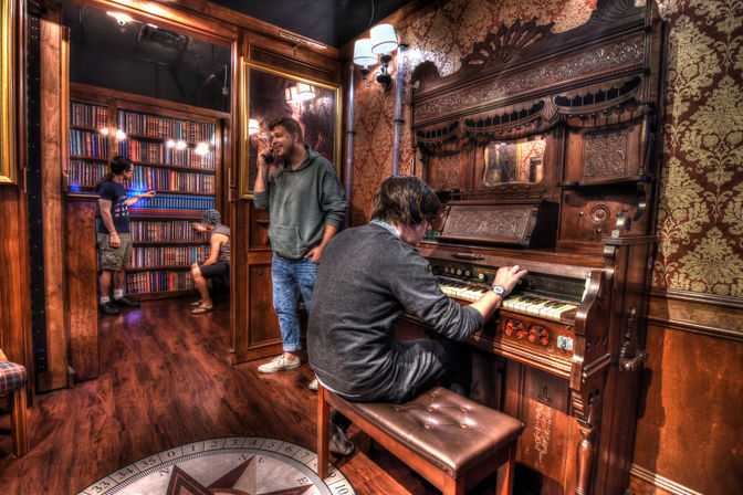Victorian-style parlor with ornate wallpaper and carved antique pump organ being played on a bench while others examine a large bookcase and a man talks on a phone near a hardwood floor with compass inlay — cozy, mysterious indoor scene.