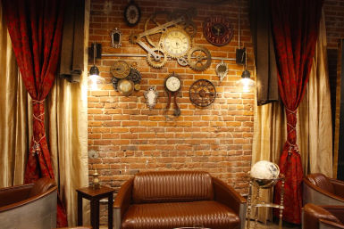 Cozy vintage loft-style lounge with exposed brick wall, brown leather sofa and armchairs, red velvet curtains, steampunk clock gears and clocks on the wall, warm pendant lights and a decorative globe.