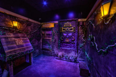 Purple-lit indoor escape room styled as a jungle temple, featuring a carved Aztec-style wall mask, weathered wooden panels, glowing lanterns, creeping vines and ornamental grasses.