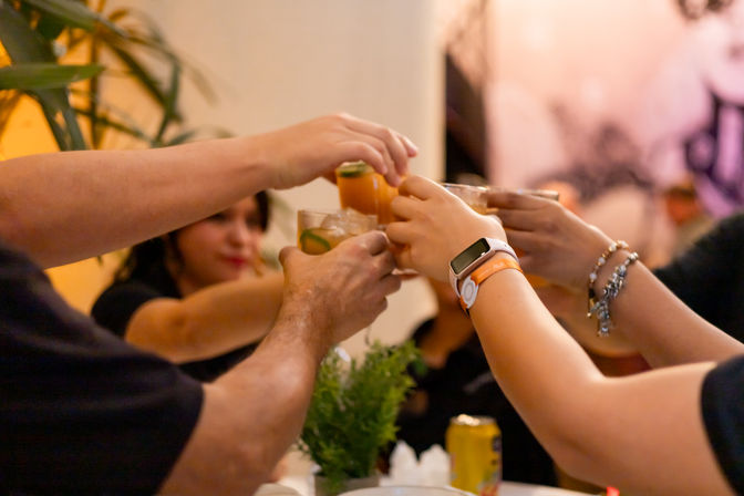 Group cheering as they clink cocktails with lime garnishes over a cozy indoor table, blurred background and visible wrist accessories.