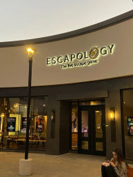 Evening outdoor shopping-center escape room entrance with glowing sign and streetlamp, glass storefront showing posters and lounge seating, and a person sitting nearby on their phone.