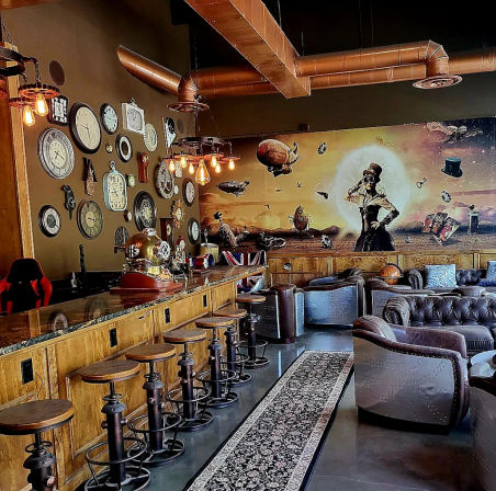Cozy steampunk lounge interior with wooden bar and swivel metal stools, wall of vintage clocks, exposed copper ductwork and Edison bulbs, large airship mural, leather tufted sofas and armchairs.