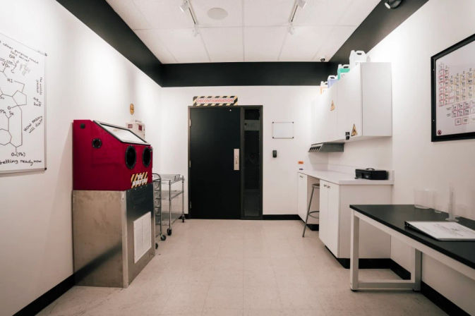 Bright white decontamination lab room with a black door labeled 'DECONTAMINATION', a red glove box unit on the left, stainless rolling cart, white cabinets and countertops on the right, and chemistry posters and a whiteboard on the walls.