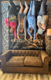 Four friends posed upside-down on a photo wall holding celebratory signs after an escape room experience in Sandy Springs, GA, above a brown tufted couch in a patterned lobby.