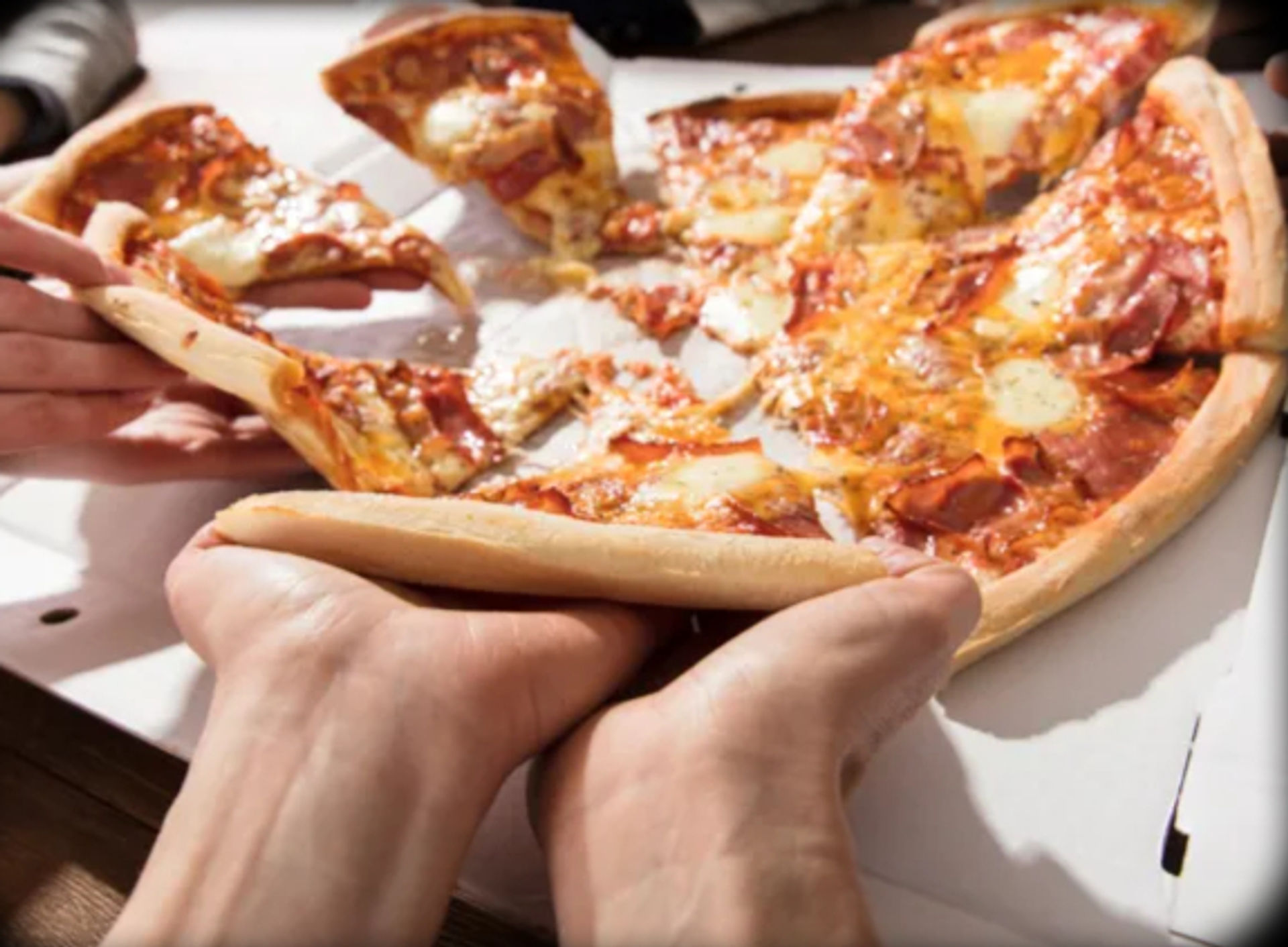 Hands pulling cheesy slices of hot pepperoni pizza from an open delivery box on a wooden table