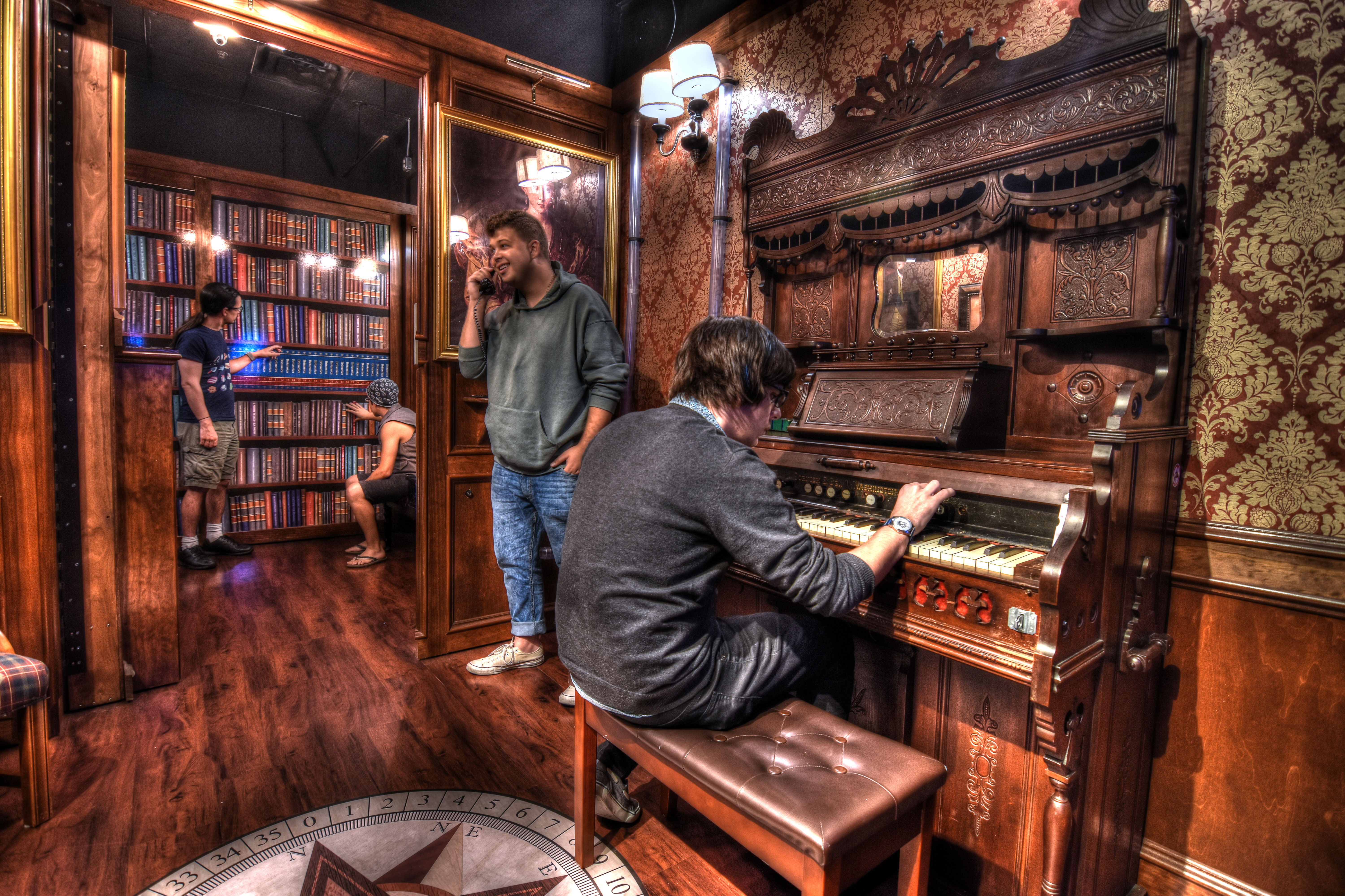 Ornate Victorian-style parlor interior with an antique pump organ, patterned wallpaper, wood-paneled bookcase and people exploring and playing music under warm lighting.