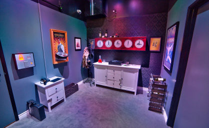 Themed escape room interior with purple mood lighting, white sideboard and nightstand, row of world clocks, vintage posters, coat stand with fur, wine rack and puzzle props on carpet.