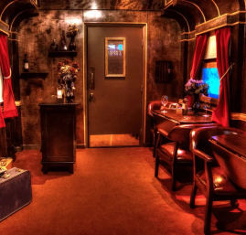 Cozy vintage train-car dining nook with wood-paneled walls, red curtains, leather chairs at a small table set with wine glasses and a bottle, floral arrangements and a suitcase by a closed door.