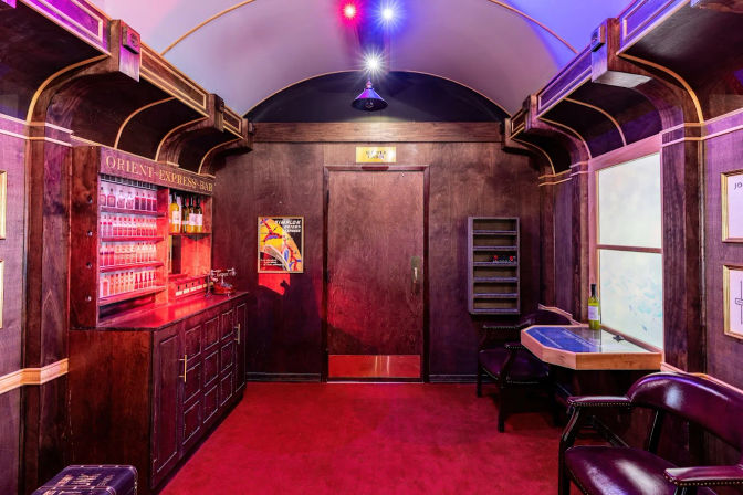 Vintage luxury train-car lounge interior with wood-paneled walls, small onboard bar, red carpet, leather seats and colorful ambient lighting.