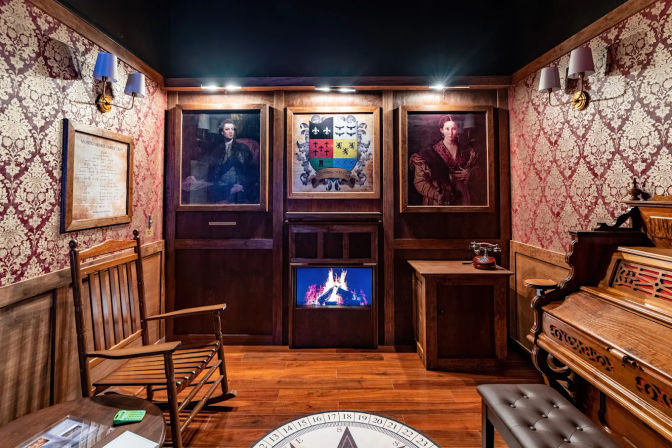 Cozy vintage parlor with dark wood paneling and red damask wallpaper, two framed oil portraits and a family crest above a glowing electric fireplace, wooden rocking chair and antique upright piano, small table with a rotary phone and warm hardwood floor.