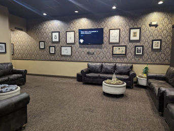 Cozy commercial lounge waiting room with dark leather sofas, patterned wallpaper, framed art, wall-mounted TV, round ottoman and carpeted floor.