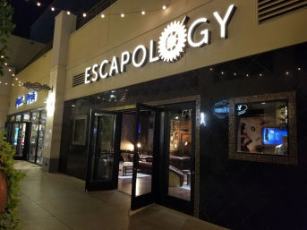 Nighttime exterior of an illuminated escape-room venue in a shopping plaza, with open glass doors revealing a cozy lounge-style interior and string lights overhead.
