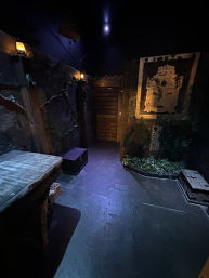 Dimly lit indoor escape-room scene evoking an ancient temple: stone-textured walls, lanterns, wooden shutter door, carved mask panel, treasure chests and jungle plants.