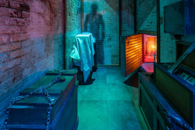 Dimly lit brick-walled cellar with teal-blue lighting, chained wooden trunk and crates, a cloth-draped object casting a humanlike shadow, and a small open hatch revealing warm orange light beyond.