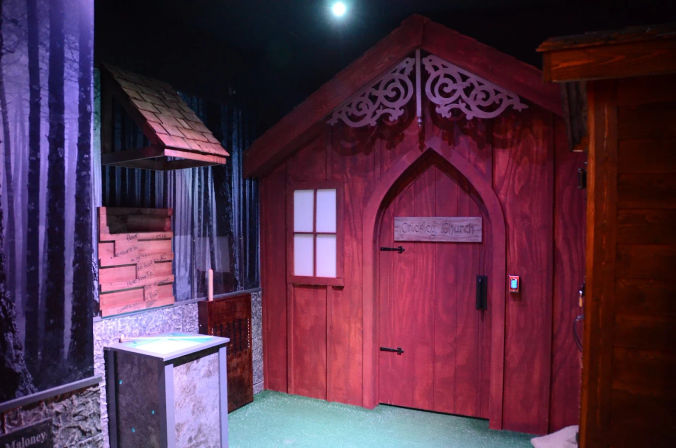 Indoor themed mini-golf scene with a red wooden cottage facade featuring an ornate gable and arched door, frosted window, small keypad, faux moonlit forest mural and green artificial turf foreground.