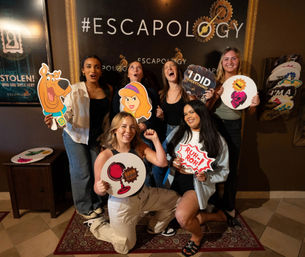 Group of six friends posing at an escape room photo wall, holding playful cutout props including Scooby-Doo and Daphne, a “Ruh‑roh!” speech bubble and an “I did everything” sign, celebrating a successful game in an indoor entertainment venue.