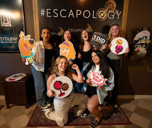 Group of six friends posing at an escape room photo wall, holding playful cutout props including Scooby-Doo and Daphne, a “Ruh‑roh!” speech bubble and an “I did everything” sign, celebrating a successful game in an indoor entertainment venue.