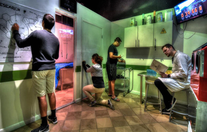 Team of four tackling a lab-themed escape room puzzle—one writes chemical formulas on a whiteboard, one kneels at a sliding door, another checks equipment at a sink, and a person in a lab coat reads a manual under cabinets with hazard symbols and a visible countdown timer.