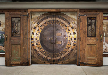 Escape-room storefront entrance featuring a giant decorative bank vault mural on double doors framed by weathered wooden shutters, set on a city sidewalk.