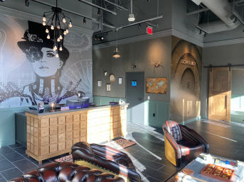 Sunlit industrial-style escape room lobby interior with a large steampunk mural of a woman, apothecary-style wooden reception counter, leather lounge chairs, pendant lights, and tiled floor.