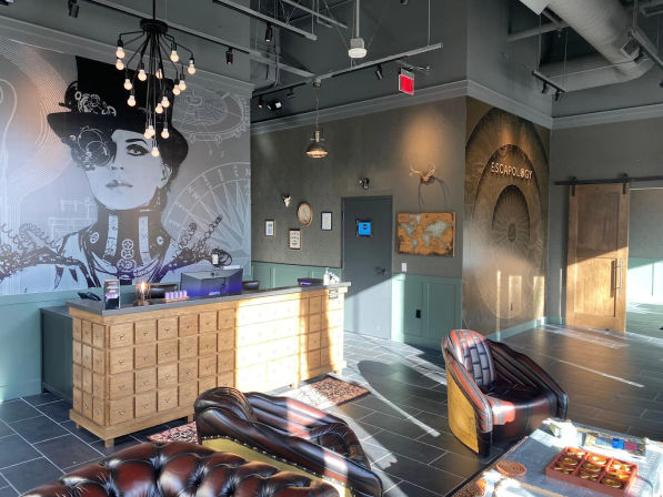 Sunlit industrial-style escape room lobby interior with a large steampunk mural of a woman, apothecary-style wooden reception counter, leather lounge chairs, pendant lights, and tiled floor.