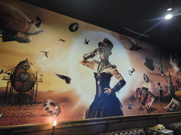 Steampunk wall mural in an indoor lounge: Victorian-dressed woman in a corset and top hat with goggles, surrounded by floating gears, a dirigible, birds, tarot cards and a retro‑futuristic capsule over a desert landscape.