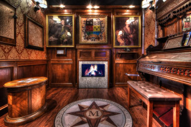 Cozy vintage wood-paneled parlor with a lit fireplace, framed portraits and coat of arms, compass-rose floor medallion, carved antique piano and tufted bench under warm ambient lighting.