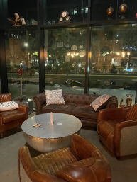 Cozy industrial-chic lounge with brown leather sofa and armchairs surrounding a riveted metal round coffee table by floor-to-ceiling windows overlooking a city street at night
