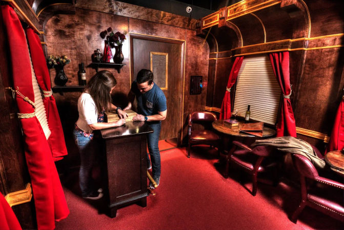 Two people huddle over a wooden podium solving a puzzle in a wood-paneled vintage parlor with red carpet, red curtains, leather chairs and small tables — cozy escape-room mystery setting.
