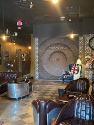 Cozy escape room lobby interior with tufted leather sofas, riveted metal chairs, a Union Jack armchair by a lamp, large compass mural and patterned wallpaper under warm pendant lighting.