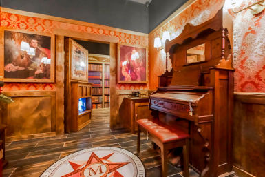 Cozy Victorian-style parlor interior with red damask wallpaper, an antique wooden upright organ and tufted bench, framed portraits and wall sconces, a doorway opening to a book-lined library, and a decorative compass medallion set into dark wood flooring.