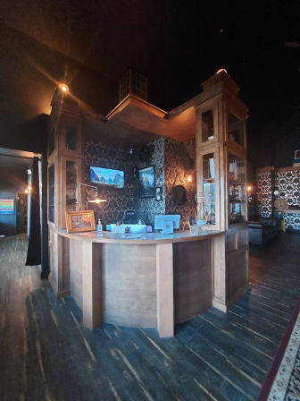 Curved wooden reception desk in a cozy, moody lobby with damask wallpaper, wall-mounted TVs, warm ambient lighting and dark hardwood floors.