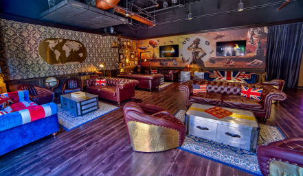 Travel-themed lounge with leather chesterfield sofas, Union Jack and US flag pillows, metal trunk coffee tables, globe, wall map and a steampunk adventure mural on hardwood floors.