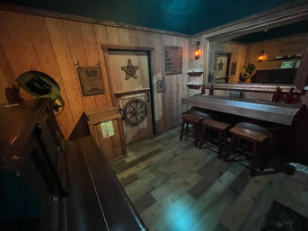 Cozy old-west saloon interior with wood-paneled walls, rustic bar counter and three stools, sheriff's star door, cowboy hats, lantern lighting and a large mirror.