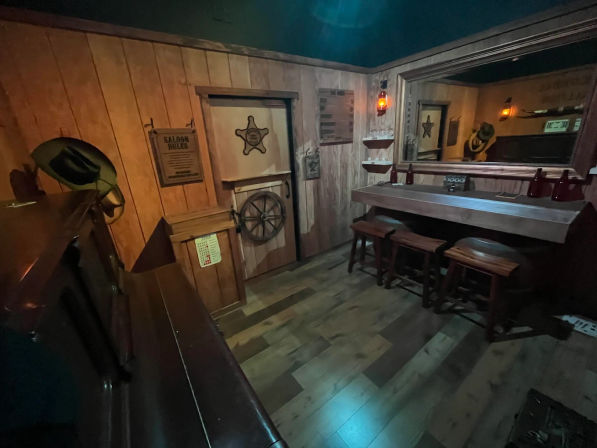 Cozy old-west saloon interior with wood-paneled walls, rustic bar counter and three stools, sheriff's star door, cowboy hats, lantern lighting and a large mirror.