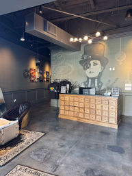Industrial-chic boutique interior with steampunk mural of a woman in a top hat and monocle, wooden apothecary-style counter with many drawers, t-shirt merch rack, leather seating, exposed ductwork, concrete floors and modern chandelier.