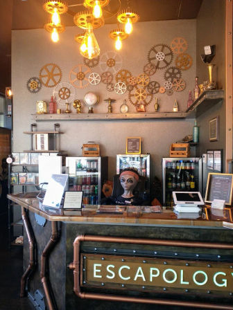 Steampunk-themed escape room reception with copper pipes, wall of gears, glowing Edison bulbs and an alien mannequin behind the counter.