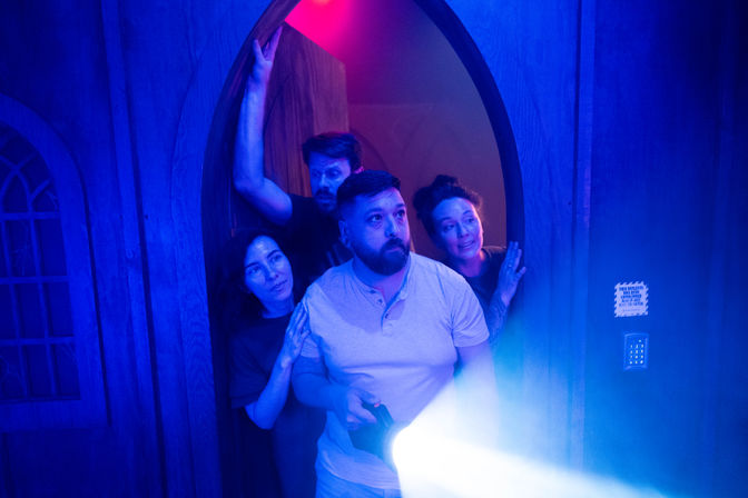 Four adults peering through an arched wooden doorway into a dim, neon-blue room as a man in front shines a flashlight—group exploring a spooky indoor escape-room or haunted attraction.