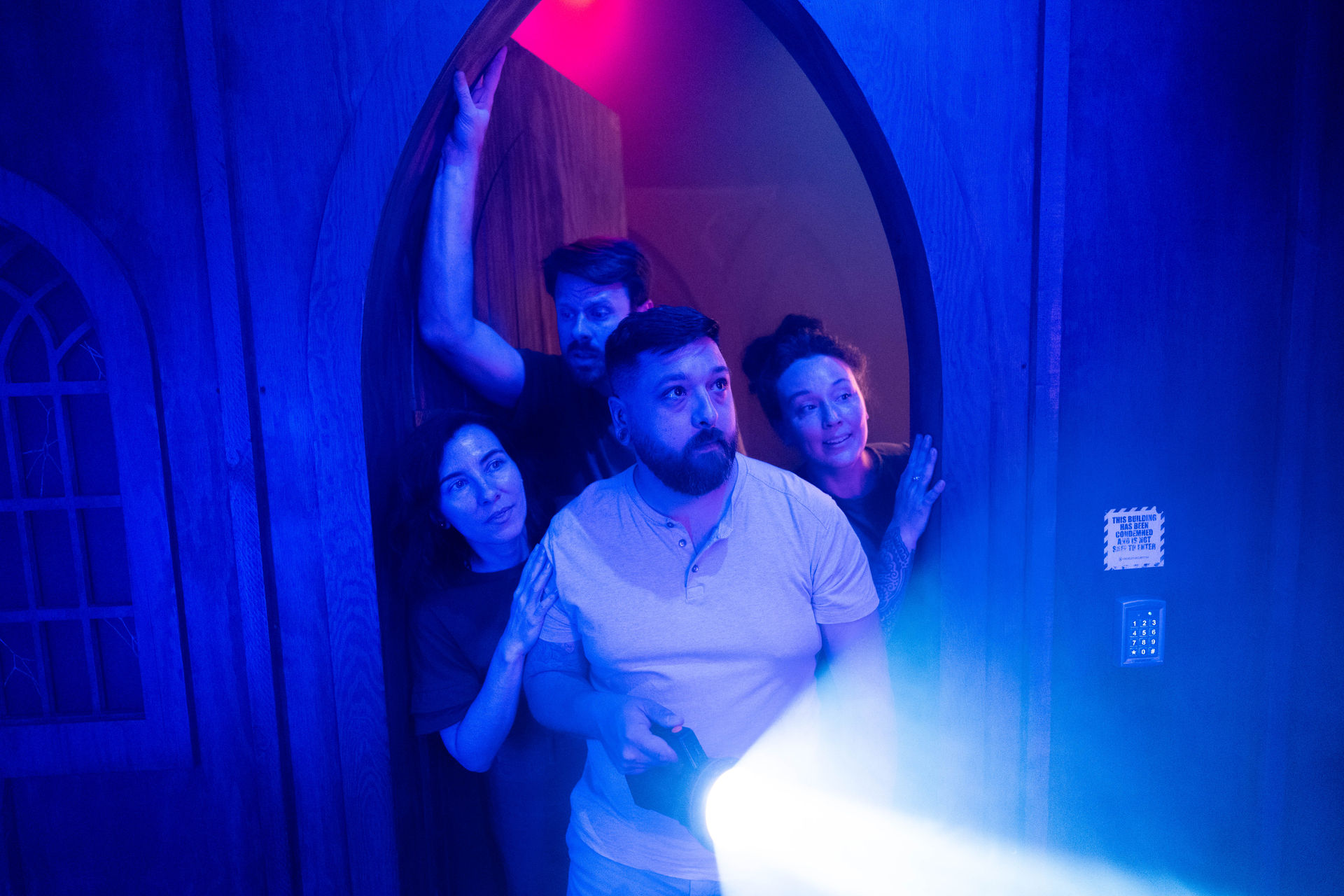 Four adults peering through an arched wooden doorway into a dim, neon-blue room as a man in front shines a flashlight—group exploring a spooky indoor escape-room or haunted attraction.