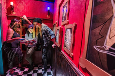 Three players huddled over an open book in a red-lit escape room styled as a spooky library with checkered floor, bookshelves, framed portraits, and glowing wall candles.
