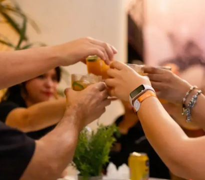Group of people clinking orange citrus cocktails in a lively indoor toast, close-up of hands and drinks with blurred guests and potted plant in background.